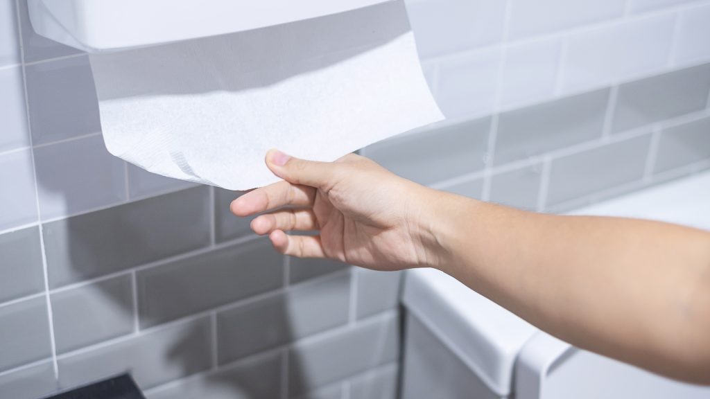 Person pulling a paper towel from a dispenser in a public washroom to dry hands — promoting hygiene and reducing germ spread.