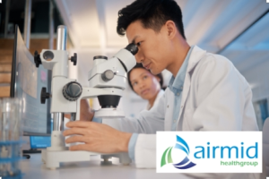 Two researchers in a bright lab operate a microscope; airmid healthgroup logo appears in the lower right corner.