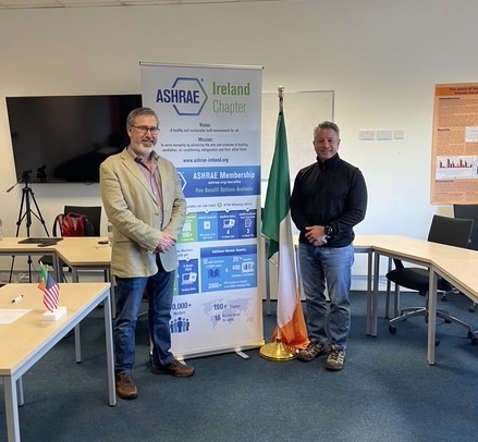 ASHRAE President Bill McQuade and ASHRAE Ireland President Howard Neville stand together at the Airmid Healthgroup facility in Dublin, with ASHRAE Ireland banners and US and Irish flags visible in the background, symbolizing the international partnership on healthy buildings and Indoor Environmental Quality