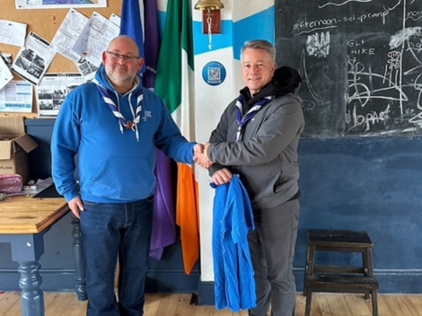 Two men in scout scarves shake hands in a room with flags, a chalkboard, and a bulletin board.