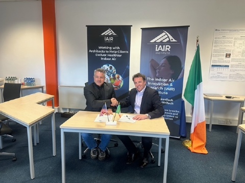 ASHRAE President Bill McQuade and Dr. John McKeon, CEO of Airmid Healthgroup, shake hands at the iAIR Institute office in Dublin, with iAIR banners and the Irish flag visible in the background, symbolizing their partnership on healthy buildings and Indoor Environmental Quality