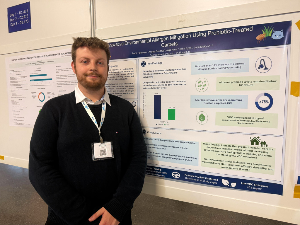 Man with conference badge standing in front of a research poster about probiotic-treated carpets at a convention.