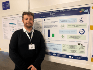 Man with conference badge standing in front of a research poster about probiotic-treated carpets at a convention.