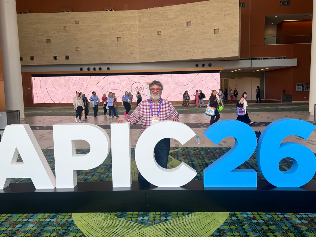 Airmid Healthgroup Scientific Consultant Matthew Hardwick standing at the large APIC26 conference sign in the Nashville convention centre atrium.